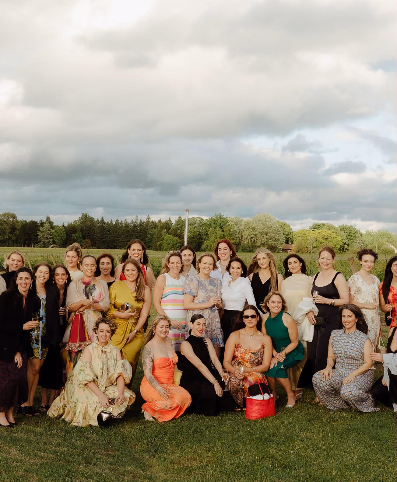 A large group of women posing outdoors on grass with trees and partly cloudy sky in the background, dressed in various colorful and stylish outfits, some standing and some sitting.