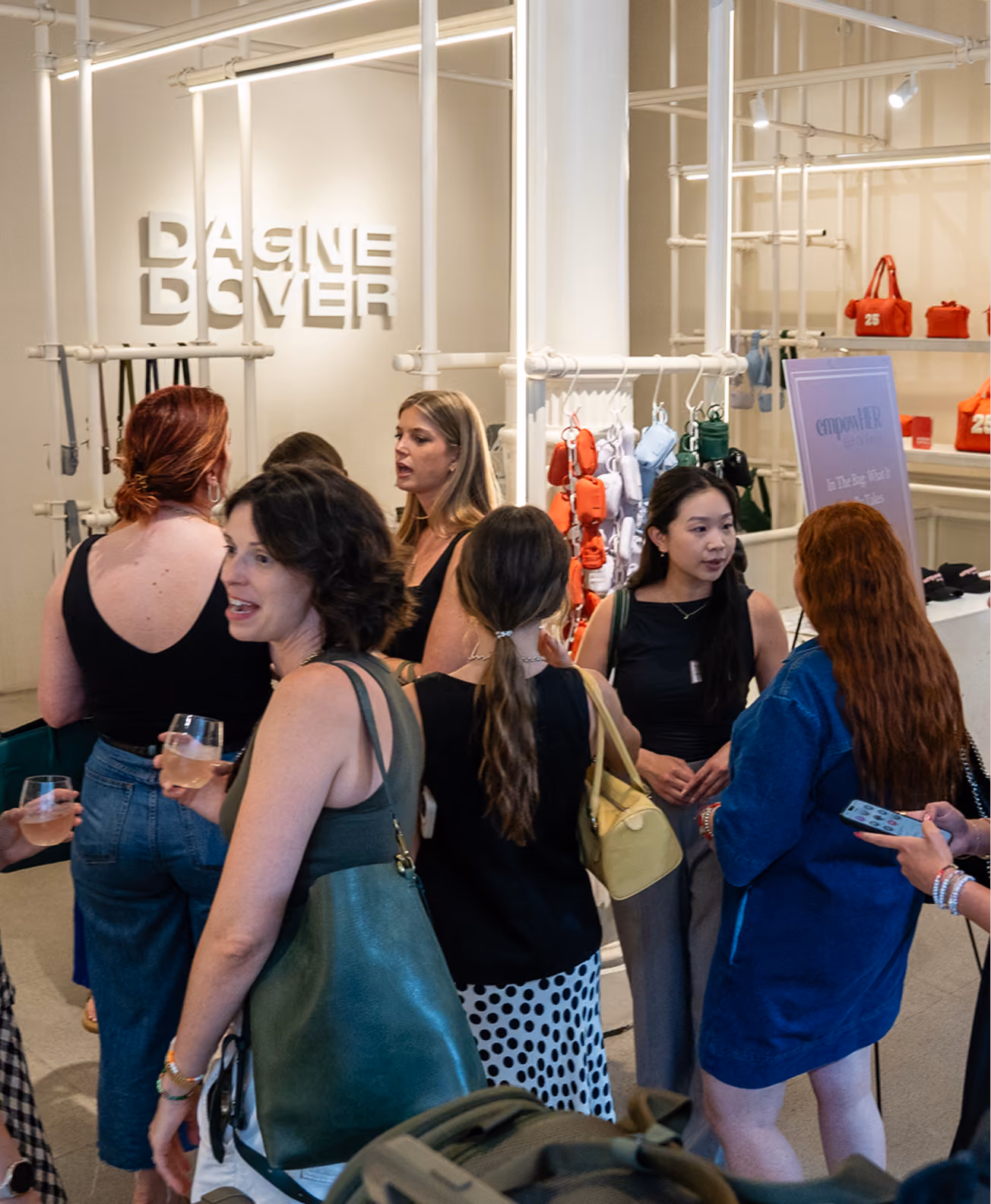 Group of women socializing inside a boutique with modern white pipe fixtures and a display of colorful handbags, some holding drinks and engaged in conversation.