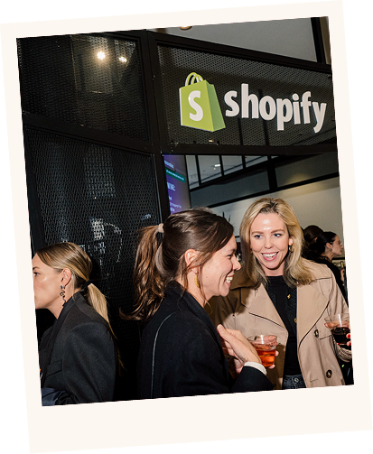 Three women socializing indoors near a black metal partition with a Shopify logo above them, two women smiling and holding drinks.