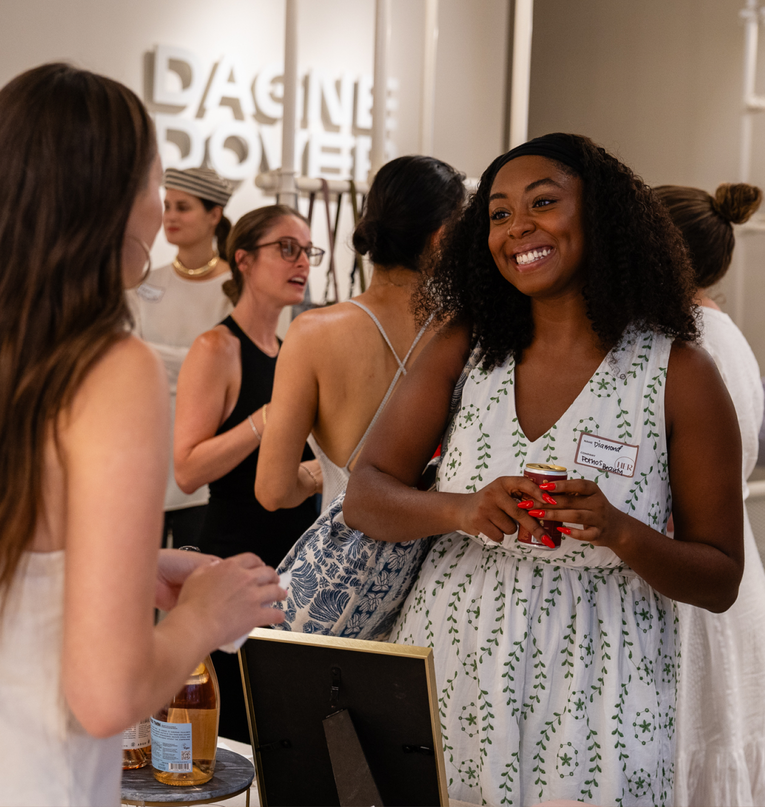 A smiling woman with curly hair wearing a white dress with green leaf patterns holds a drink can while talking to another person at a social gathering with several people in the background.