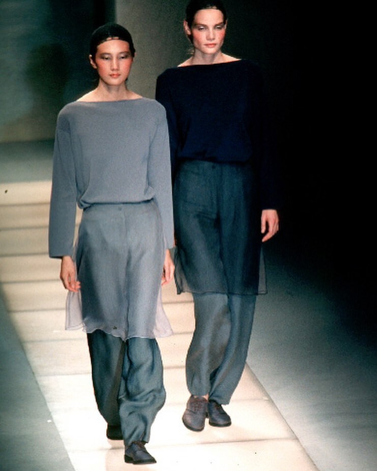 Two female models walking on a runway wearing sheer tunics over loose pants and dark shoes.
