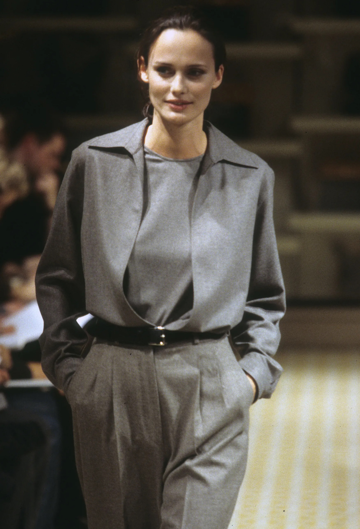 Model walking on runway wearing a grey collared jacket, matching high-waisted trousers, and hands in pockets.