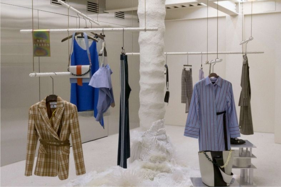 Modern minimalist clothing display with hanging garments including a plaid blazer, blue dress, striped shirt, and various skirts in a white room.
