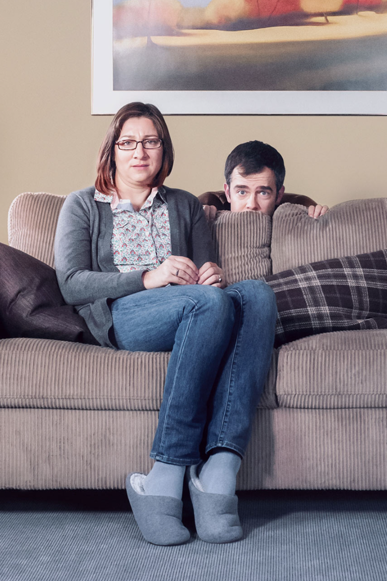 Couple sitting on sofa while man hides behind cushion in living room, Zoocasa campaign.