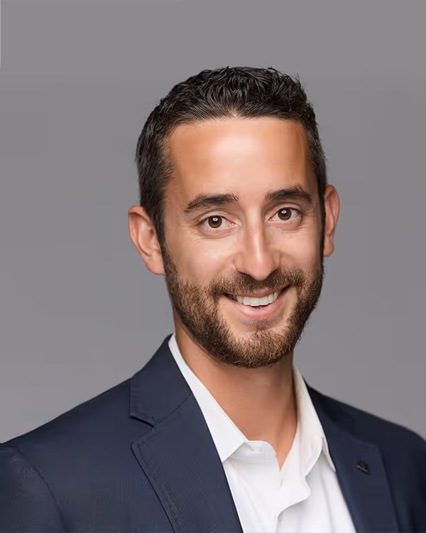 Smiling man with short curly hair and beard wearing a navy blazer and white shirt against a gray background.