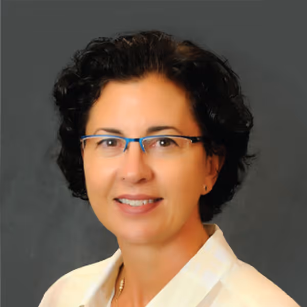 Woman with short curly dark hair, wearing blue-rimmed glasses and a white collared shirt, smiling against a gray background.