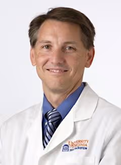 Smiling man wearing a white lab coat with University of Virginia Health System logo and a blue shirt with striped tie.