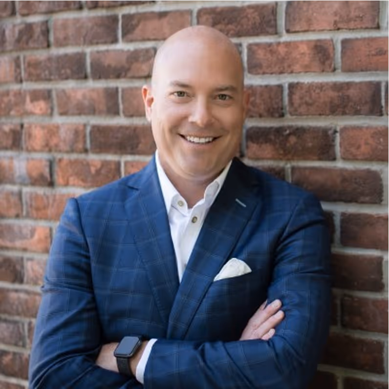 Smiling bald man in a blue checkered suit jacket and white shirt standing with arms crossed against a brick wall.