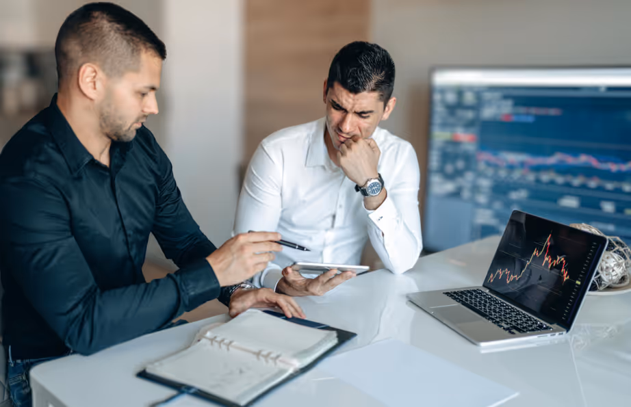Two colleagues in the office with a laptob showing graphs and a notebook on the desk discussing work issues.