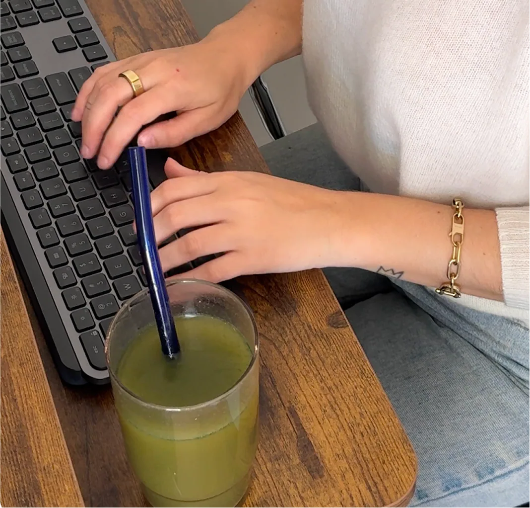 A lady typing using a keyboard with a drink standing next to her on a table
