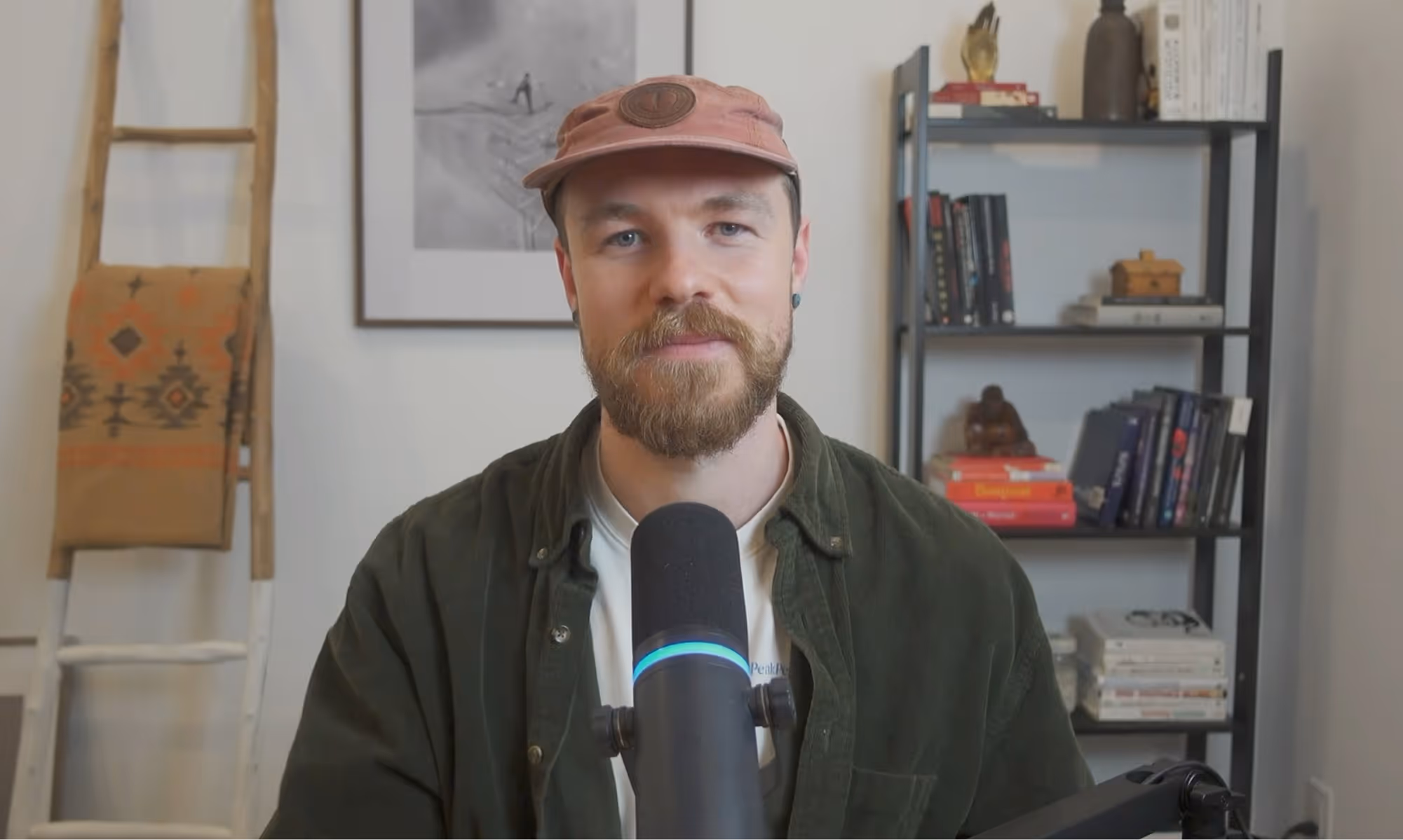 Bearded man wearing a pink cap and dark green shirt speaking into a microphone with shelves and a ladder in the background.