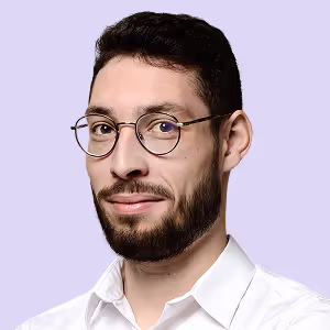 Portrait d'un homme avec des lunettes rondes, barbe et chemise blanche devant un fond violet clair.