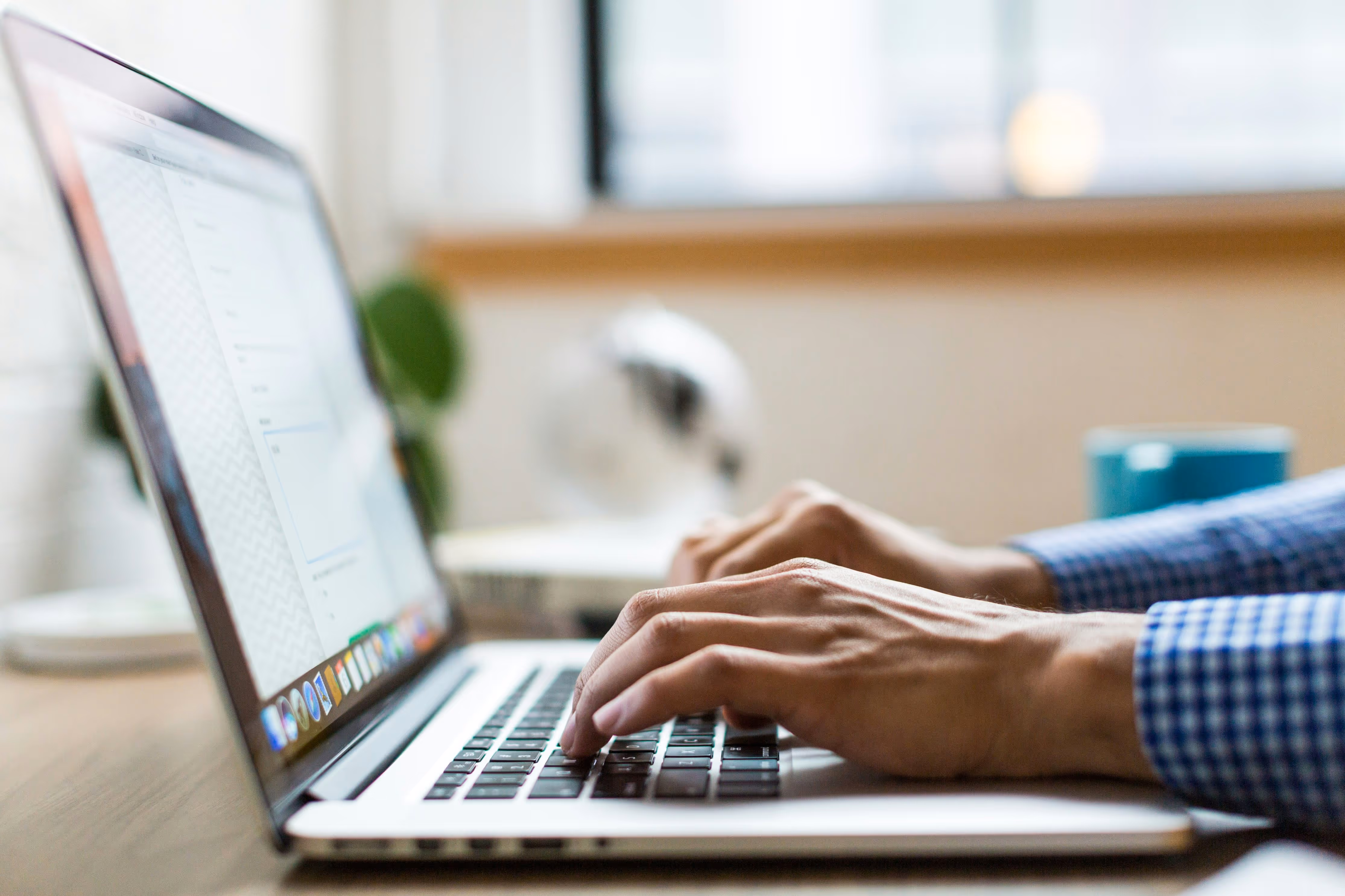 Person typing on a laptop keyboard with a blurred background of a window and desk items.
