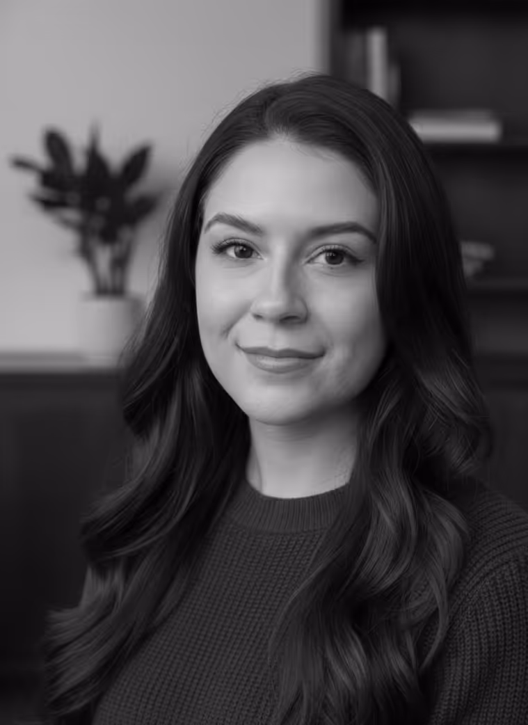 Black and white portrait of a woman with long wavy hair wearing a knitted sweater, smiling gently with a blurred indoor background.