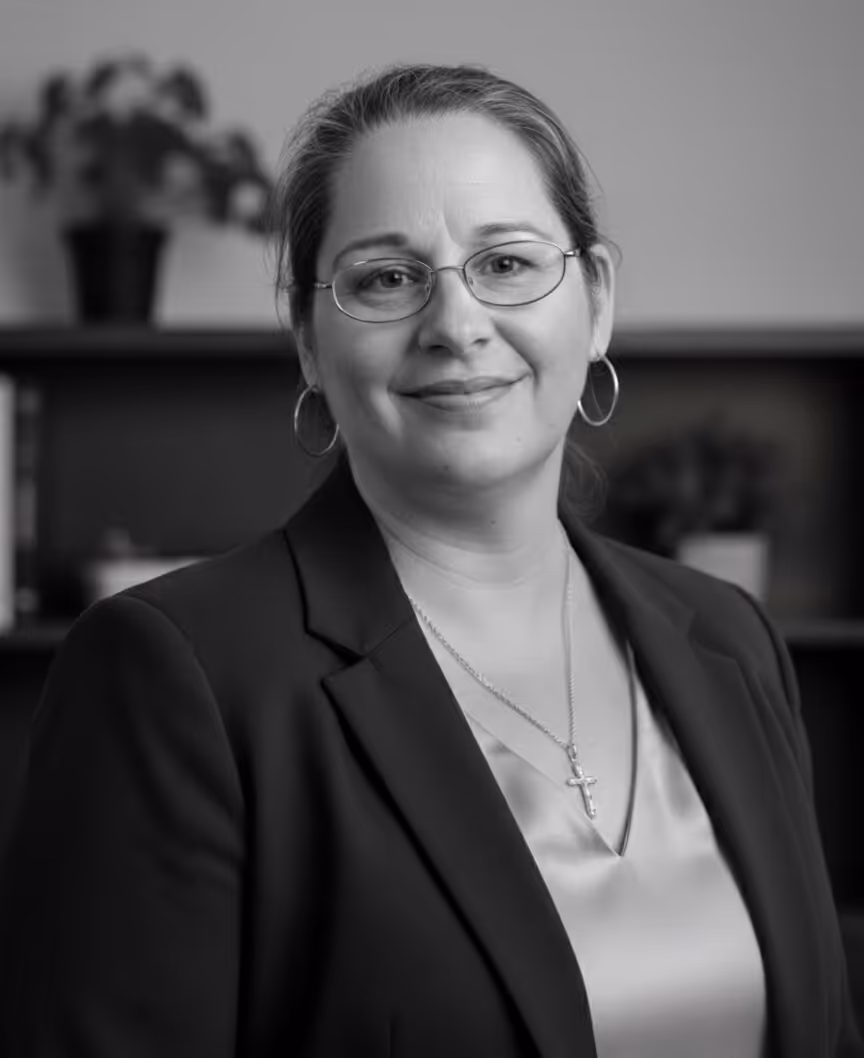 Smiling woman wearing glasses, hoop earrings, and a cross necklace, dressed in a blazer and blouse, with a blurred background.