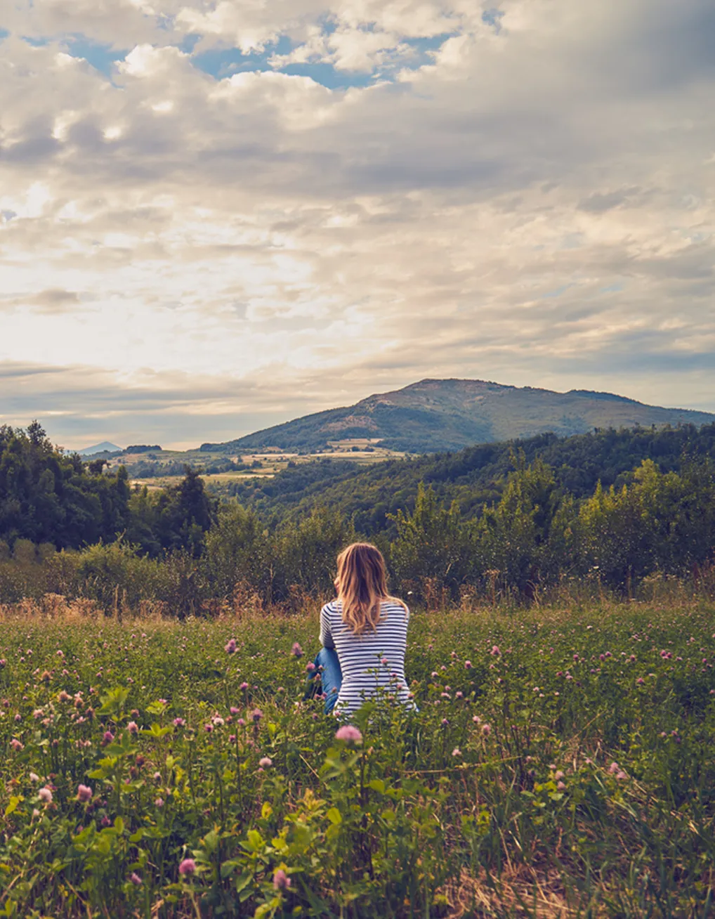 Person with long hair and striped shirt sitting in a wildflower meadow, facing forested hills under a cloudy sky.