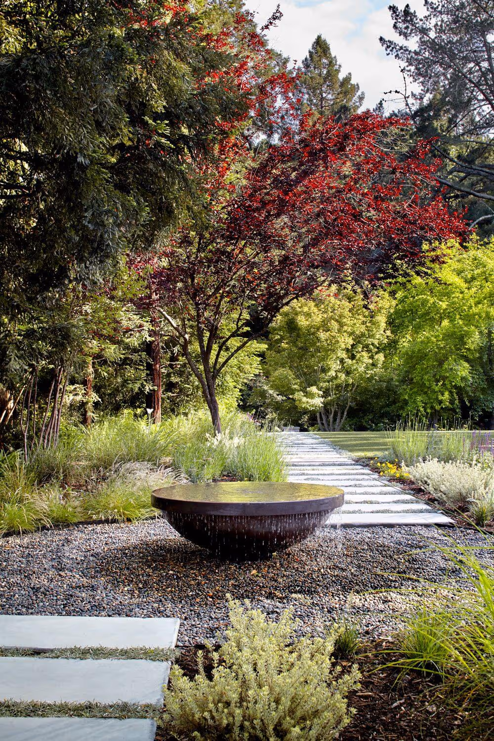 Modern circular water feature surrounded by gravel in a landscaped garden with green and red-leaved trees and a stone pathway.