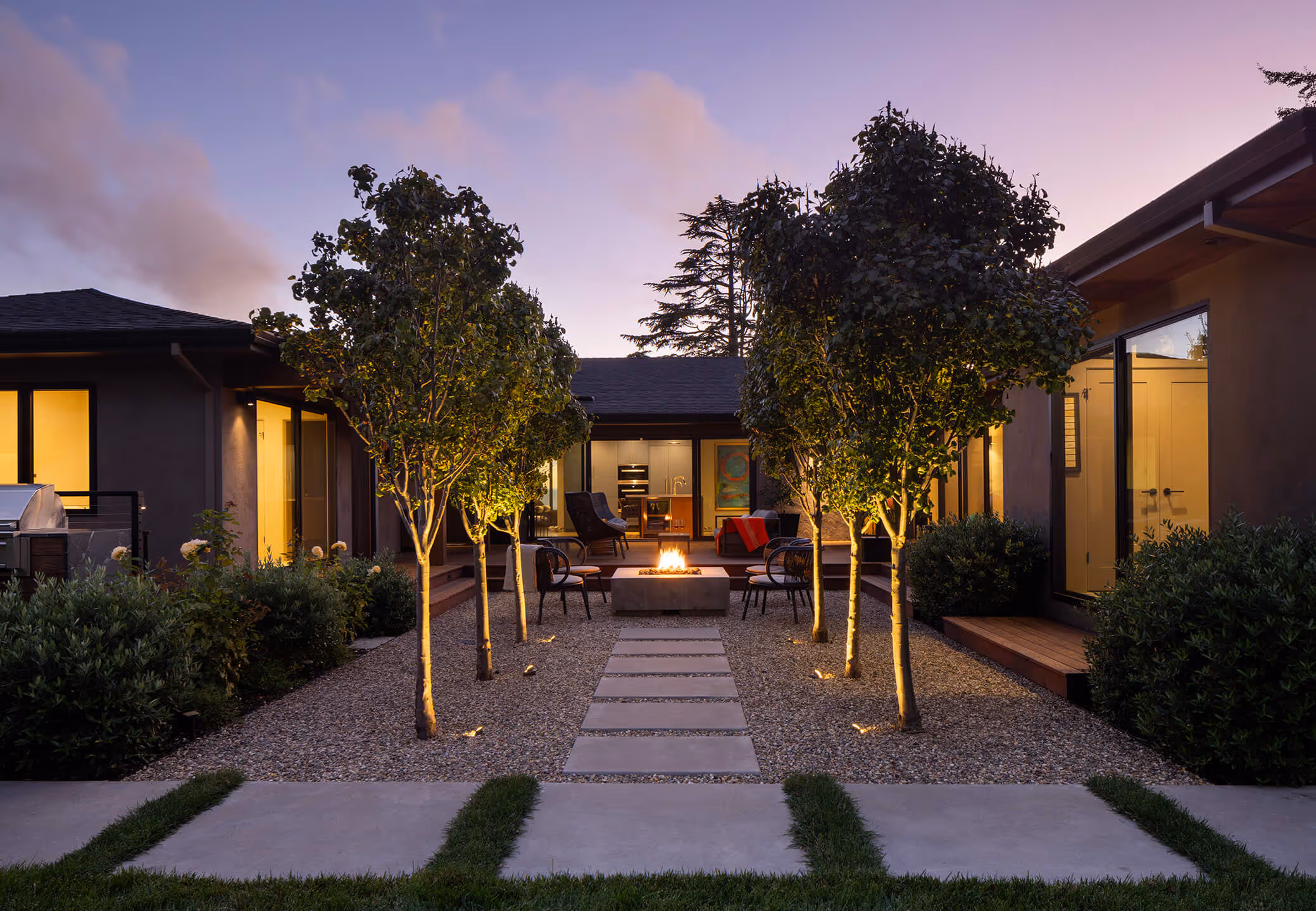 Modern backyard patio at dusk with lit fire pit, seating area, and pathway flanked by small illuminated trees and bushes.