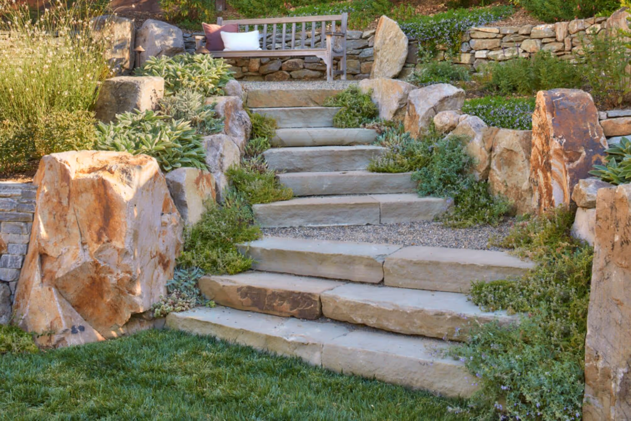Stone staircase with greenery on both sides leading to a wooden bench with pillows in a landscaped garden.