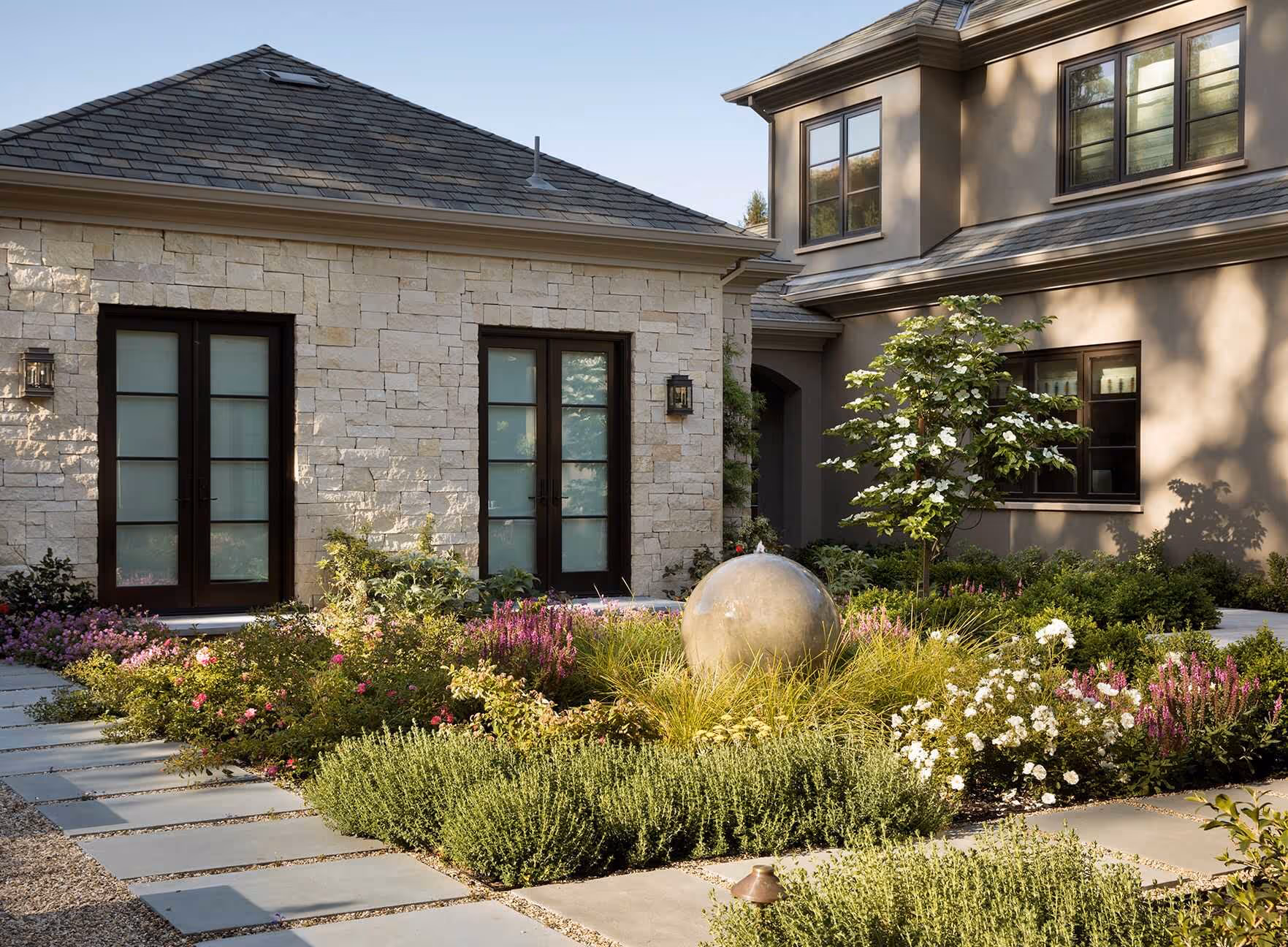 Modern courtyard garden with stone walkway, lush green shrubs, flowering plants, and a large spherical stone water feature in front of a beige stone and stucco house.