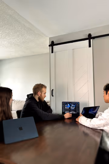 Three people sitting around a table engaged in a discussion with laptops in a modern room with a white sliding barn door.