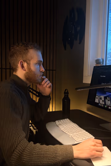 Taylen Sather, owner and founder of Aryze Design sitting at desk, focused on a computer screen with keyboard and mouse in dimly lit room.
