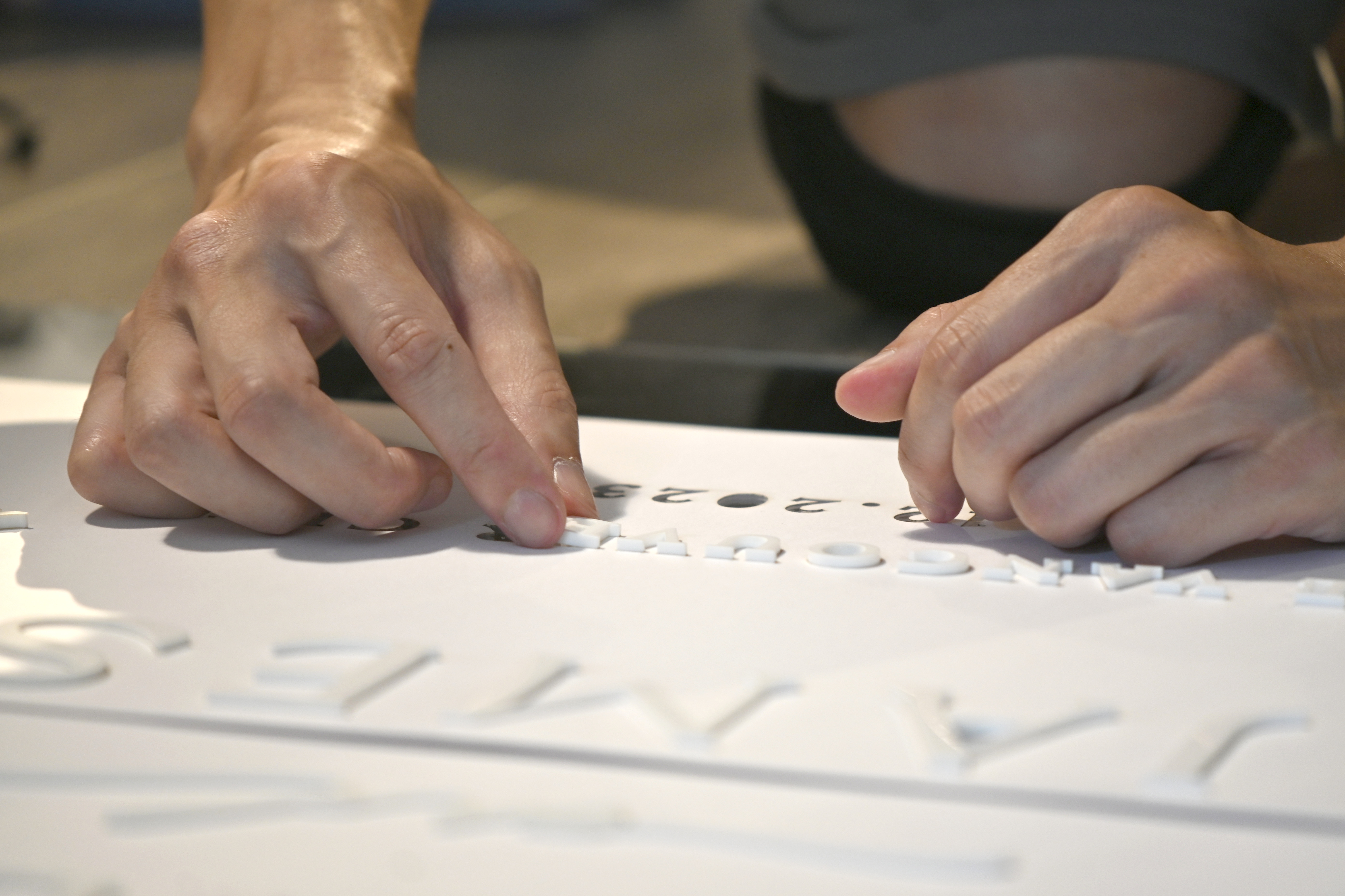 A person's hands arranging raised white letters on a white surface with partially visible black printed text.