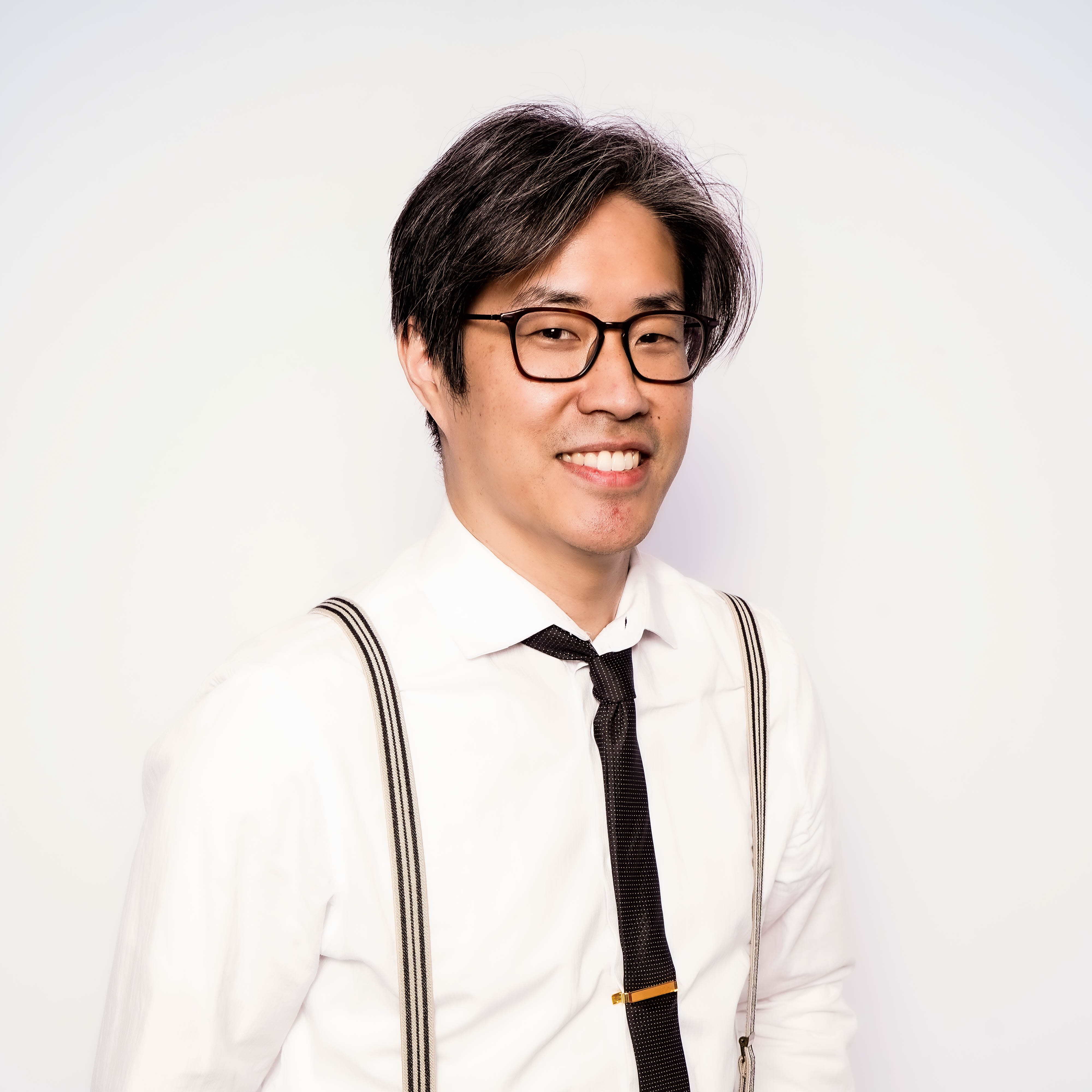 Image of Ray, founder of Printed by Ray. Smiling man with glasses wearing a white shirt, black tie with tie clip, and striped suspenders against a white background.