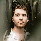 Portrait of a young man with light brown hair and beard, looking forward through green leaves.