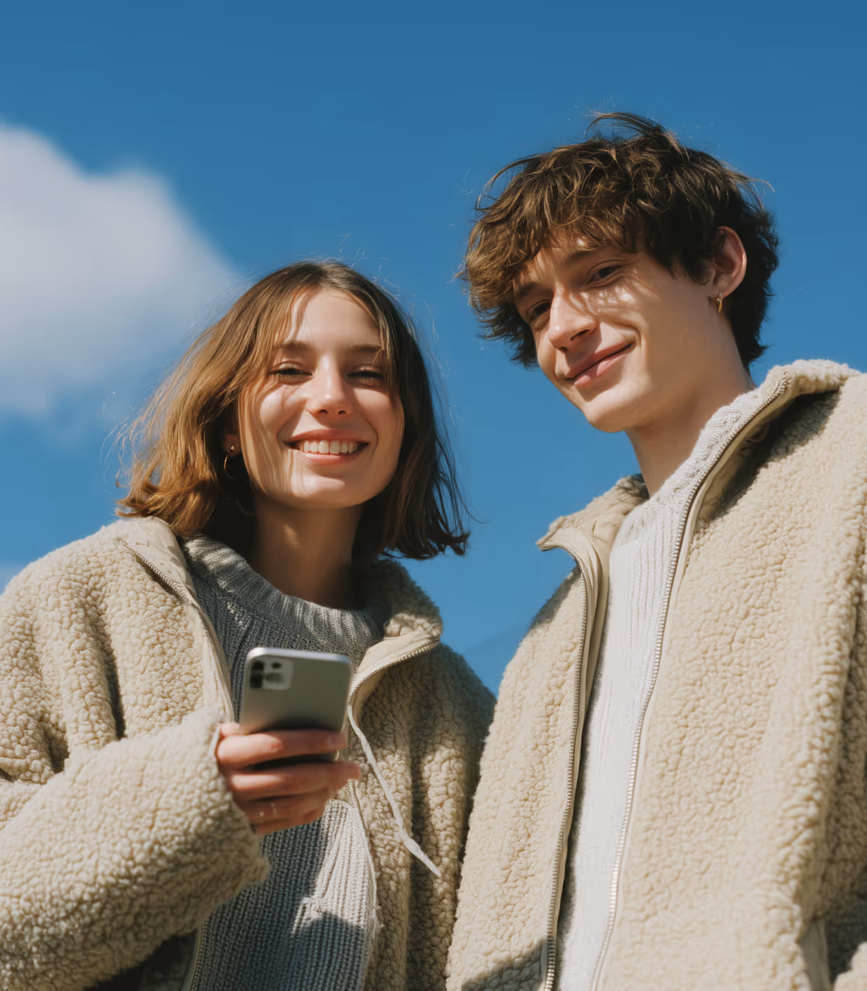 Smiling young woman and man wearing beige fleece jackets, outdoors under a clear blue sky; the woman holds a smartphone.