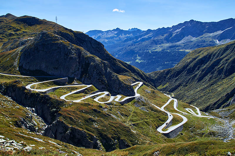 Serpentinenstraße auf einer grünen Berglandschaft mit hohen Bergen im Hintergrund unter blauem Himmel.
