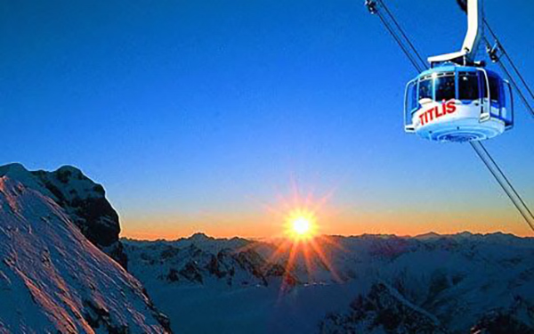 Sunset over snow-covered mountain peaks with a Titlis cable car suspended on the right.