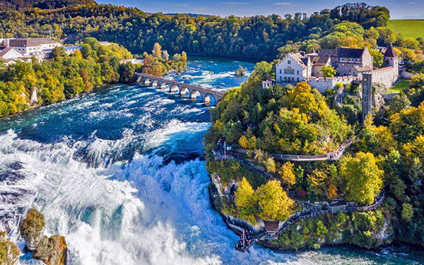 Aerial view of a wide waterfall flowing beside a green forested cliff with a castle on top and a stone bridge crossing over the river.