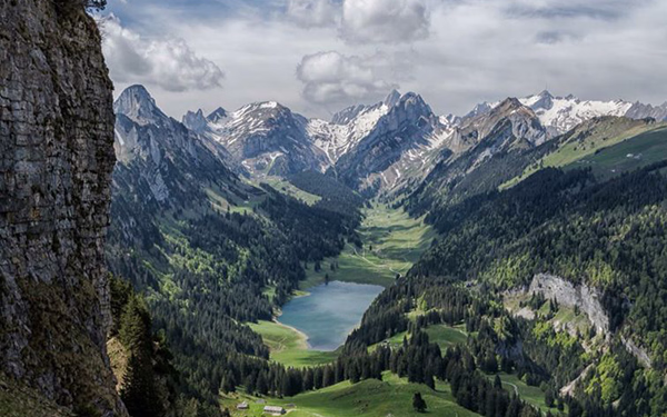 Mountain valley with a narrow lake surrounded by green forests and rugged snowy peaks under a cloudy sky.