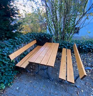 Holztisch mit zwei Bänken auf gepflastertem Boden, umgeben von Büschen und einem Baum.
