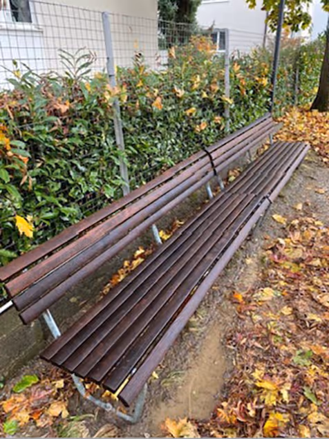 Lange braune Holzbänke im Freien, umgeben von herbstlichen Blättern und einem Zaun mit grünen Pflanzen im Hintergrund.