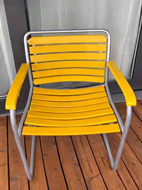 Gelber Stuhl mit Armlehnen und grauem Metallgestell auf Holzterrasse vor Glasfenster.