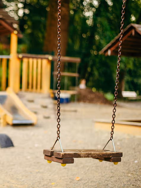 Leere Holzschaukel an Ketten auf einem Spielplatz mit unscharfem Hintergrund aus Klettergerüst und Bäumen.