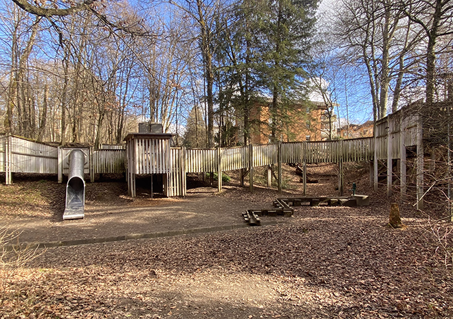 Spielplatz im Wald mit Holzkonstruktion, Rutsche, Brücke und Sandkasten, umgeben von kahlen Bäumen.