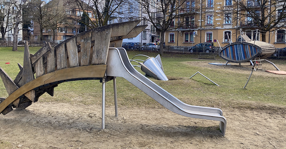 Spielplatz mit einer Rutsche in Form eines Drachens aus Holz und Metall, umgeben von Gras und Wohnhäusern im Hintergrund.