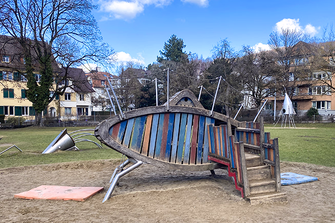 Bunter Fisch-Klettergerüst-Spielplatz im Freien mit Häusern und Bäumen im Hintergrund.