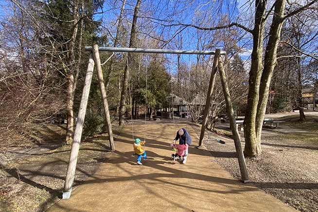 Ein Erwachsener hilft einem Kind, das auf einer Schaukel im sonnigen Park sitzt, während ein weiteres Kind auf der benachbarten Schaukel spielt.