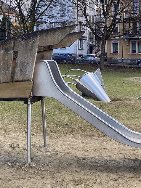 Leere Holzschaukel hängt an Ketten auf einem Spielplatz mit Rutsche im Hintergrund.