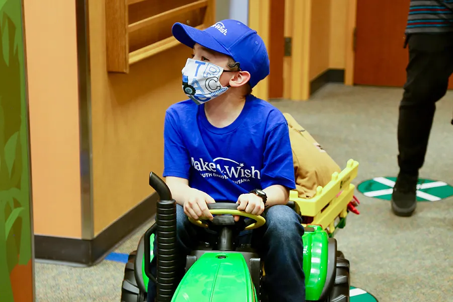 Child on green toy tractor wearing blue cap.