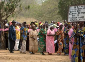 A line of women in bright African-print dresses and headscarves marching in a dusty outdoor protest; a sign to the right.