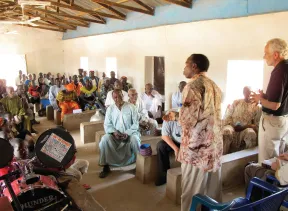 Crowded, sunlit hall with wooden beams as a man in a patterned shirt speaks to a seated, attentive audience.