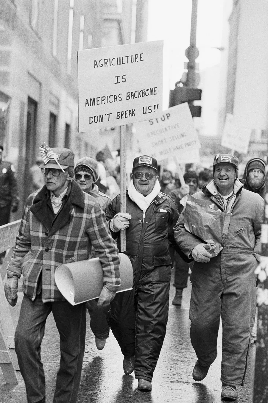 Black-and-white street protest; man in cap and scarf carries a sign reading "AGRICULTURE IS AMERICA'S BACKBONE, DON'T BREAK US!!"