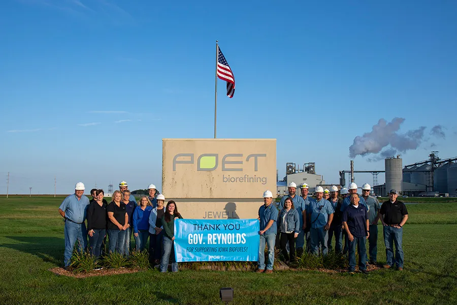 Group of workers in hard hats stand before a POET biorefining sign, holding a blue banner as American flag flies overhead.