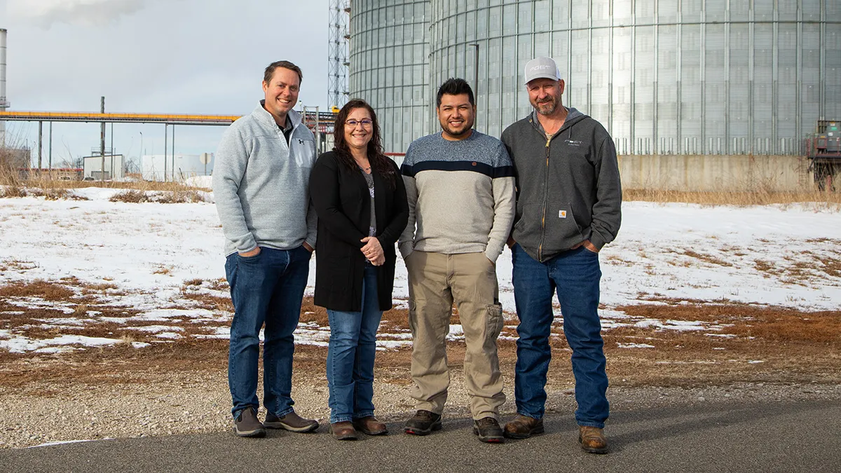 Four people pose outside grain silos.