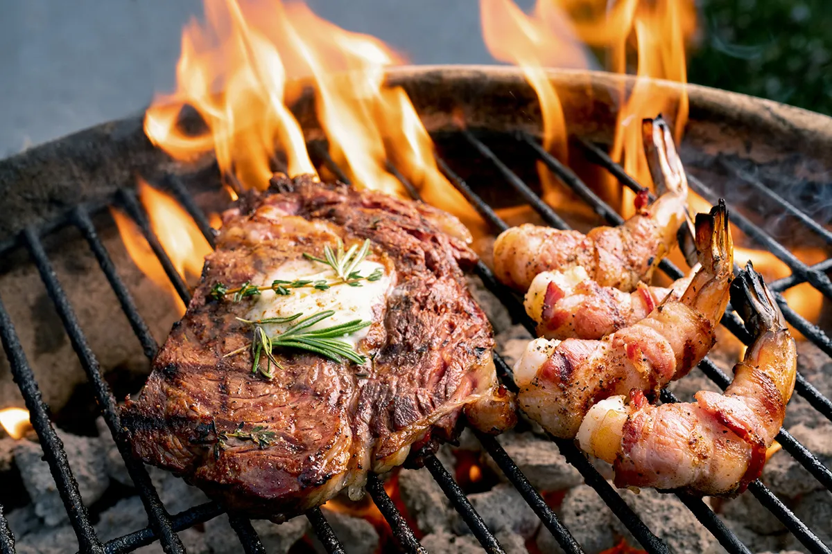 Juicy grilled steak with rosemary butter on one side, bacon-wrapped shrimp skewers nearby, flames licking the charcoal grill.